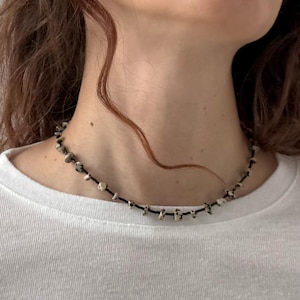 May include: A black cord necklace with small, irregularly shaped, off-white beads. The necklace is worn against a white t-shirt. The beads have dark spots, creating a speckled appearance. The necklace is a choker style.