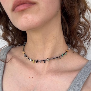 May include: A choker necklace featuring a thin black cord strung with small, irregularly shaped, multi-colored gemstone chips. The stones include shades of yellow, green, blue, white, and dark gray, spaced evenly along the length of the cord.