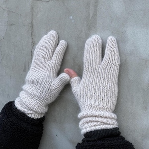 Puede incluir: Un par de guantes de punto beige claro con un diseño único de tres dedos. Los guantes tienen puños acanalados y se usan sobre una manga negra. El fondo es una pared gris lisa.
