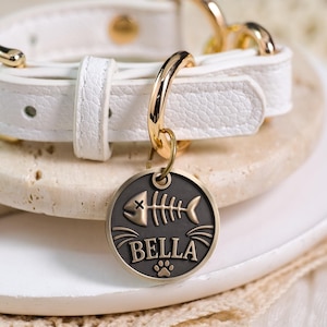 Peut inclure: Un collier blanc texturé avec des ferrures en métal doré et une médaille circulaire en métal foncé. La médaille présente un motif gravé d'arête de poisson, le nom BELLA en lettres capitales et une petite icône de patte en bas au centre.