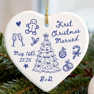 May include: A heart-shaped ceramic ornament with blue illustrations and text. The ornament says "First Christmas Married" and "May 16th 2025". A snowman with a black hat and scarf is in the background.