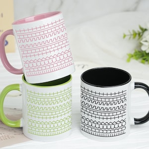 Peut inclure: Trois mugs en céramique avec des extérieurs blancs et des motifs. Un mug a un intérieur et une anse roses, un autre a une anse verte, et le troisième a un intérieur et une anse noirs. Les mugs sont empilés.