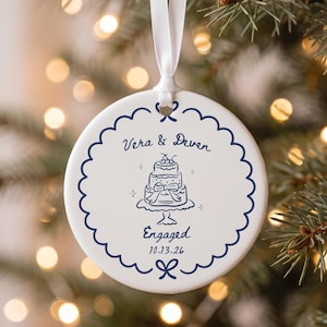 Könnte beinhalten: Ein rundes, weißes Keramikornament mit einem dunkelblauen, gewellten Rand, das an einem weißen Band hängt. In der Mitte befindet sich eine Strichzeichnung einer Torte, der Text "Vera & Deven", "Engaged" und das Datum "10.13.26" in Dunkelblau.