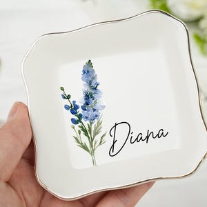 以下が含まれることがあります： 金色の縁と水彩画の花柄が施された白いセラミック製の小物入れ。青い花と筆記体で「Diana」という名前が特徴です。ジュエリーや小物を入れる装飾品です。