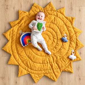 May include: A mustard yellow quilted play mat shaped like a sun with pointed rays. A baby lies on the mat holding a small book with a green leaf illustration. Attached to the mat are a rainbow toy with pom-poms and two small plush clouds.
