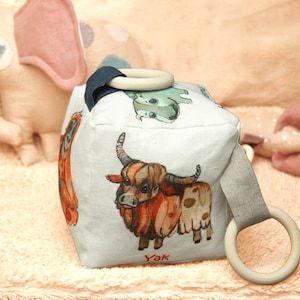 May include: A white fabric cube with a variety of animal illustrations, including a yak, a monkey, and an elephant. The cube has two wooden rings attached to it, making it a fun and interactive toy for babies and toddlers.