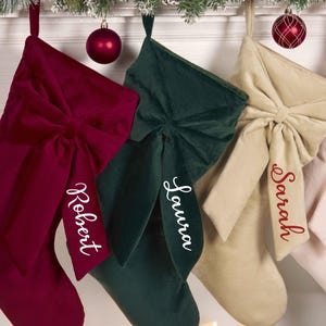 Peut inclure: Quatre bas de Noël en velours bordeaux, vert, beige et rose, chacun avec un grand nœud et un nom brodé en rouge. Les bas sont suspendus à une cheminée décorée avec des ornements et de la verdure.