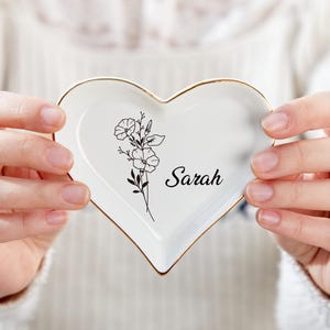 Könnte beinhalten: Eine weiße, herzförmige Schale aus Keramik mit goldenem Rand. Die Schale zeigt ein schwarz-weißes Blumenmuster und den Namen "Sarah" in Schwarz.