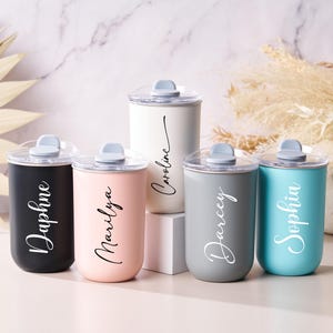 May include: A collection of five personalized tumblers in various colors: black, pink, white, gray, and teal. Each tumbler features a clear lid, a light gray flip-top, and a name in elegant script. The tumblers are cylindrical and appear to be made of metal.