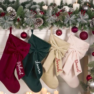 Puede incluir: Cuatro calcetines navideños de terciopelo en burdeos, verde, beige y rosa, cada uno con un lazo grande y un nombre bordado en rojo. Los calcetines cuelgan de una repisa decorada con adornos y vegetación.