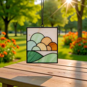 May include: A stained glass panel with a black frame, depicting a landscape with rolling hills. The hills are in shades of green, yellow, and orange. The panel is hanging from a black cord, and the background is a sunny outdoor scene.