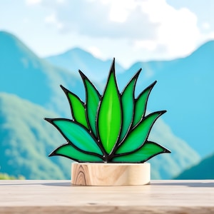 Puede incluir: Una escultura de planta de agave de vidrieras con hojas verdes vibrantes y contornos negros, montada sobre una base de madera natural. La obra de arte se encuentra sobre un fondo de montañas y un cielo azul brillante.