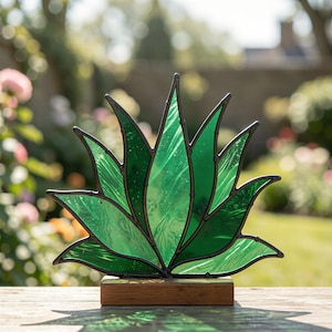Könnte beinhalten: Eine Agaven-Skulptur aus Buntglas mit mehreren spitz zulaufenden grünen Glasblättern und dunklen Lötstellen, montiert auf einem rechteckigen Holzsockel. Das Glas hat eine texturierte, durchscheinende Optik, die Licht durchlässt.
