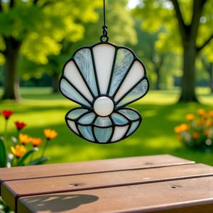 Puede incluir: Un atrapasoles de concha de vidrio manchado con un borde negro, paneles de vidrio blanco y azul-verdoso, y una cuenta central en forma de perla. El atrapasoles cuelga contra un fondo borroso de hierba verde y árboles.