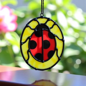 Peut inclure: Un capteur de soleil en vitrail représentant une coccinelle. La coccinelle a un corps rouge avec des points noirs et une bordure jaune. Le capteur de soleil est suspendu par une chaîne noire. L'arrière-plan est flou avec du feuillage vert et une fleur rose.