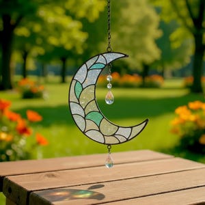 Op de afbeelding: Een gebrandschilderde halve maan zonnevanger met een zwart metalen frame. De maan is gesegmenteerd met tinten groen, blauw en wit glas. Een ketting en kristallen druppels hangen aan de maan en reflecteren zonlicht. De zonnevanger wordt buiten tentoongesteld.