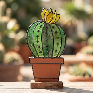 Op de afbeelding: Een glas-in-lood cactussculptuur met een rond groen lichaam met donkere metalen lijnen en kleine zwarte kralen, bekroond met een felgele bloem. De cactus staat in een terracotta-kleurige pot op een rechthoekige houten voet.