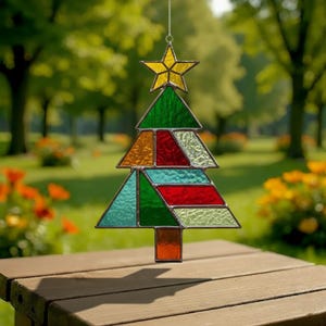 May include: A stained glass Christmas tree suncatcher with a yellow star on top. The tree is composed of various colored glass pieces, including green, red, orange, and teal. It hangs from a silver chain and is displayed outdoors.
