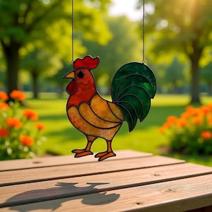 May include: A stained glass rooster suncatcher with a red comb and wattle, yellow body, and green tail feathers. The decorative rooster is hanging outside on a sunny day, with a blurred green background of trees and flowers.