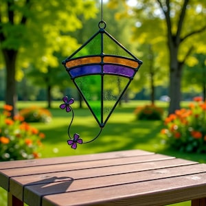 May include: A stained glass kite-shaped suncatcher with a black metal frame. The kite features green, orange, purple, and blue glass panels. A black metal stem with purple flower accents extends from the kite. The suncatcher hangs in front of a blurred green background.