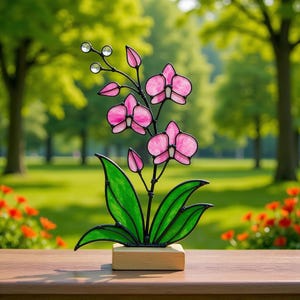 Puede incluir: Una escultura de orquídea de vidrieras con pétalos de flores rosas, hojas verdes y detalles de cristal transparente. La escultura está montada sobre una pequeña base de madera. El fondo presenta un paisaje verde borroso.