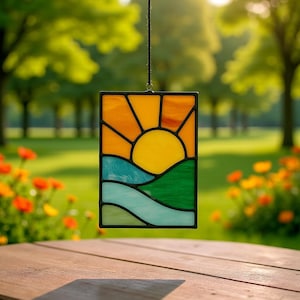 May include: A rectangular stained glass panel depicting a sunrise over rolling hills and a river. The sun is yellow, with orange rays. The hills are green, and the river is blue. The panel is framed in black and hangs from a black cord.