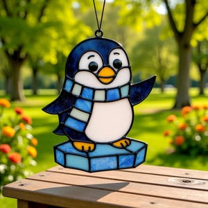 May include: A stained glass penguin ornament with a blue and white scarf and a yellow beak. The penguin is standing on a blue ice floe. The ornament is hanging from a black string. The background is a blurred outdoor scene.