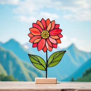 May include: A stained glass flower sculpture with a vibrant red, orange, and yellow flower head, green leaves, and a black stem. The flower is mounted on a small wooden base. The background features a blurred mountain range and blue sky.