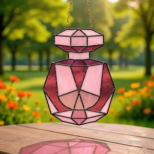 May include: A stained glass perfume bottle suncatcher in shades of pink and red, with a geometric design. The suncatcher is suspended by a gold chain, with a blurred background of green trees and flowers.