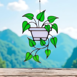 Pode incluir: Decoração de planta suspensa em vitral com um vaso branco e folhas verdes. As folhas são de cor verde vibrante e o vaso é branco. A planta é suspensa por uma corda preta. O fundo mostra uma cordilheira desfocada.