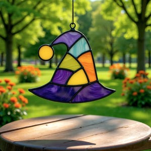 Peut inclure: Un attrape-soleil en vitrail en forme de chapeau de sorcière avec une boule jaune et une variété de couleurs, dont le violet, l'orange, le jaune et le sarcelle. Le chapeau est suspendu devant un fond vert flou avec des arbres et des fleurs.