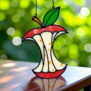 May include: A stained glass apple core suncatcher. The red and white glass is outlined in black, with a green leaf and orange stem. The suncatcher is suspended from a black string.