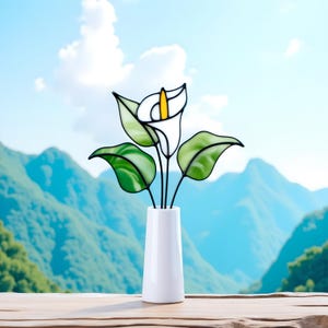 May include: A white ceramic vase holding a stained glass calla lily. The lily has white petals with a yellow center and green leaves. The background features a mountain range and a blue sky.