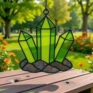 May include: A stained glass crystal suncatcher with a black metal frame. The crystal is composed of three green glass shards, with a brown base. The suncatcher is hanging from a black metal ring. The background is a blurred outdoor scene.