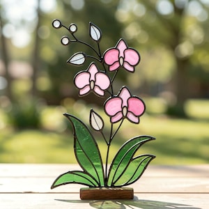 Pode incluir: Uma escultura de orquídea em vitral com três flores cor-de-rosa de centros brancos, folhas verdes e pequenos botões brancos sobre uma estrutura de metal preto. A peça está montada numa base de madeira retangular e exibe um trabalho detalhado.