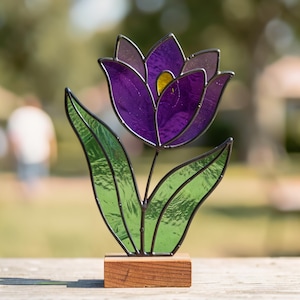 Op de afbeelding: Een glas-in-lood tulp ornament met dieppaarse bloemblaadjes, een klein geel hart, twee getextureerde groene bladeren en zwarte metalen soldeerlijnen. De glazen bloem is bevestigd op een rechthoekige houten voet voor presentatie.