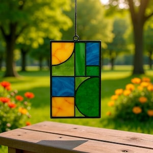 May include: A rectangular stained glass panel with a black border, featuring geometric shapes in yellow, green, and blue. The panel is suspended by a black cord. The background is a blurred outdoor scene with trees and flowers.