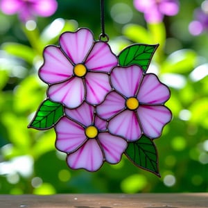 Puede incluir: Un atrapasoles floral de vidrieras con tres flores rosas, centros amarillos y hojas verdes. Las flores tienen un tono rosa claro con acentos rosas más oscuros. La pieza está diseñada para colgar y se encuentra sobre un fondo verde borroso.