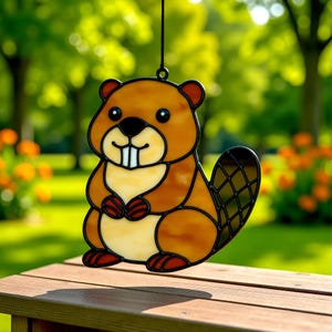 May include: A stained glass beaver suncatcher with a brown body, black tail, and cream-colored belly. The beaver has a friendly expression with two large white teeth. The suncatcher is hanging in front of a blurred green background.
