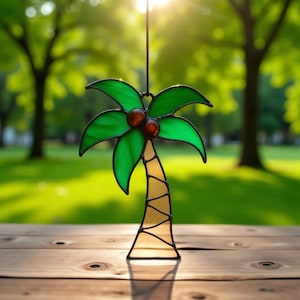 Puede incluir: Un adorno de vidriera con forma de palmera con hojas verdes y dos cocos marrones. El tronco es de color amarillo claro. El adorno cuelga frente a un fondo verde borroso.