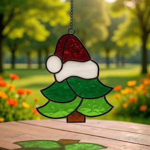 May include: Stained glass Christmas tree ornament with a Santa hat design. The ornament features a red hat, white trim, and green tree shape. It hangs from a black chain. The background is a blurred outdoor scene with trees and flowers.