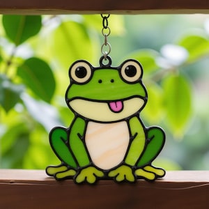 May include: A stained glass frog ornament. The frog is green with black outlines, a pink tongue, and large eyes. It hangs from a metal chain. The background is a blurred view of green leaves and foliage.