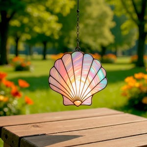 Peut inclure: Un coquillage en vitrail suspendu, aux couleurs irisées rose, bleu et jaune. La coquille est suspendue par une chaîne, avec un arrière-plan vert flou d'arbres et de fleurs. La coquille est suspendue au-dessus d'une table en bois.