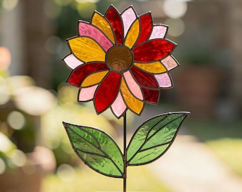 May include: A stained glass flower sculpture featuring a central brown disc surrounded by petals in shades of red, yellow, and light pink. The flower is supported by a thin metal stem with two green glass leaves, all mounted on a rectangular wooden base.