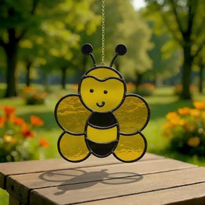 May include: A stained glass bee suncatcher with a smiling face, yellow wings, and a black and yellow body. The bee is suspended from a gold chain. The background is a blurred outdoor scene with trees and flowers.