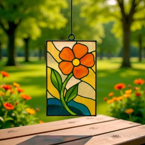 Op de afbeelding: Een rechthoekig glas-in-loodpaneel met een gestileerde oranje bloem met een gele kern, groene bladeren en een blauwe basis. Het paneel wordt opgehangen door een zwart koord. De achtergrond is een vage buitenscène met bomen en gras.