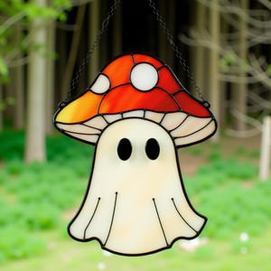 Könnte beinhalten: Ein Glasmalerei-Dekoration mit einem Pilz und einem Geist. Die Pilzkappe ist rot, orange und gelb mit weißen Punkten. Der Geist ist weiß mit schwarzen ovalen Augen. Das Stück ist schwarz umrandet und hängt an einer Kette.