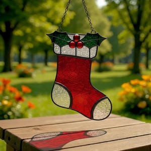 Könnte beinhalten: Ein Weihnachtsstrumpf-Dekoration aus Glas. Der rote Strumpf hat eine weiße Manschette und eine transparente Spitze und Ferse. Die Oberseite zeigt grüne Stechpalmenblätter und rote Beeren. Er hängt an einer Kette.