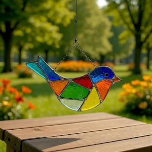 Puede incluir: Un atrapasoles de pájaro de vidrieras de colores. El pájaro está compuesto por varias formas geométricas en tonos de azul, verde, naranja, rojo y amarillo. Cuelga de una cadena, con un fondo verde borroso de árboles y flores.