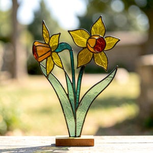 May include: A stained glass sculpture of two daffodils featuring yellow and orange petals with green glass stems and leaves. The floral piece is mounted on a rectangular wooden base, showcasing intricate dark metal soldering between the glass segments.